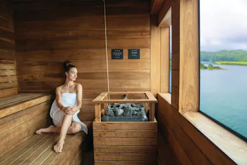 Woman in towel seated in wooden sauna beside stone heater, large window overlooking lake