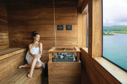 Woman in towel seated in wooden sauna beside stone heater, large window overlooking lake