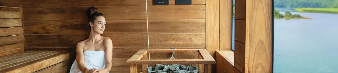 Woman in towel seated in wooden sauna beside stone heater, large window overlooking lake