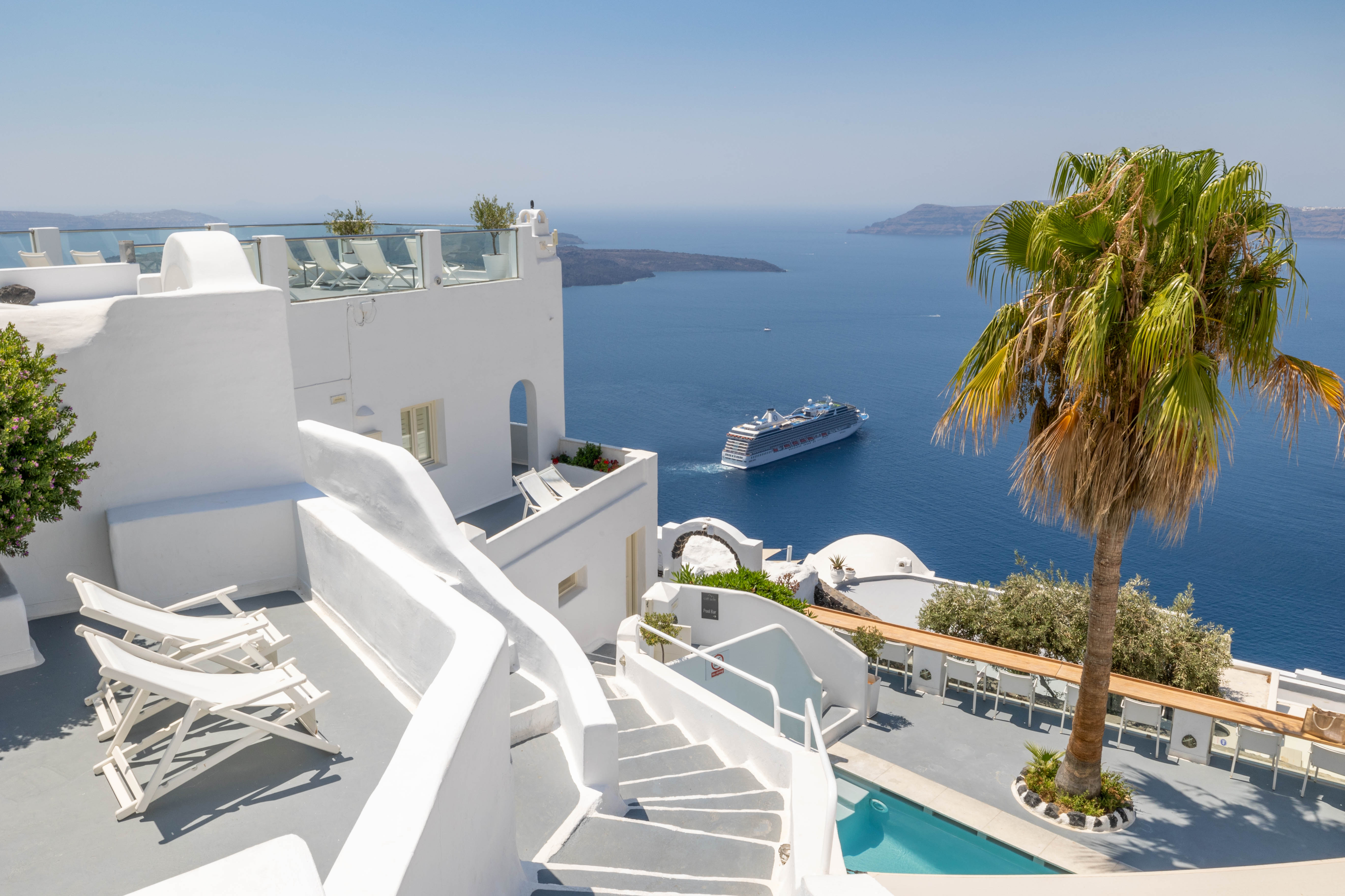 White Santorini terraces, stairs and pool overlooking Aegean Sea with cruise ship and palm tree.