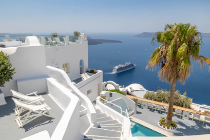 White Santorini terraces, stairs and pool overlooking Aegean Sea with cruise ship and palm tree.