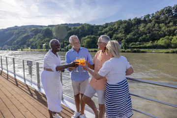Four older friends toasting with orange cocktails on a riverboat deck with tree-lined hills.