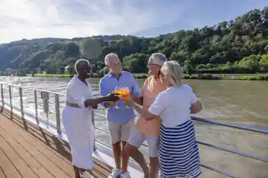 Four older friends toasting with orange cocktails on a riverboat deck with tree-lined hills.