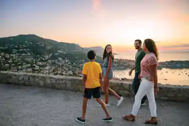 Smiling family of four strolling at seaside overlook at sunset, harbor and hills below