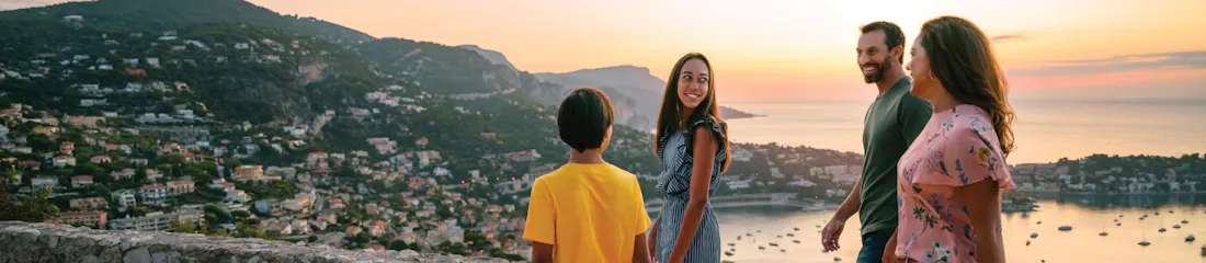Smiling family of four strolling at seaside overlook at sunset, harbor and hills below