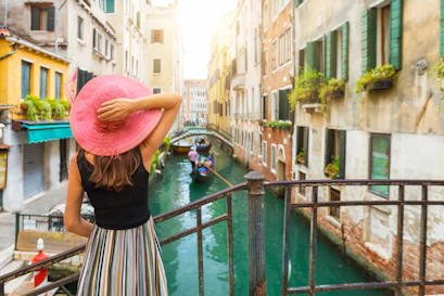 Back-view woman in pink sunhat overlooking Venice canal with gondola from an iron bridge