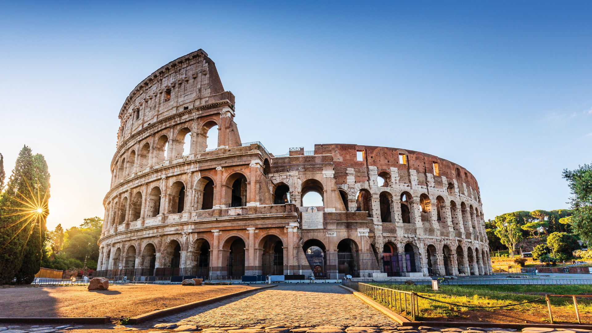 Colosseum in Rome at sunrise, sunburst behind trees, cobbled path in foreground