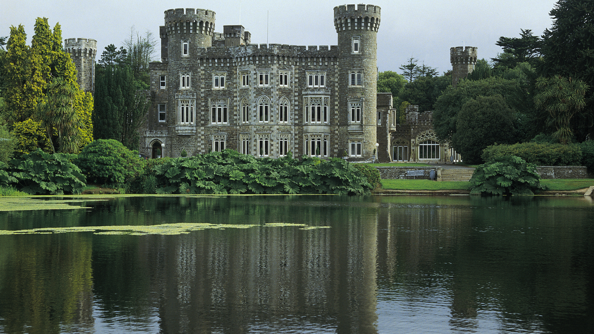 Historic stone crenellated castle with round towers and many windows, reflected in a leafy ponda