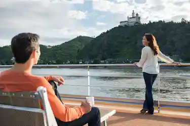 Woman leans on river cruise railing viewing hilltop castle while man sits with coffee