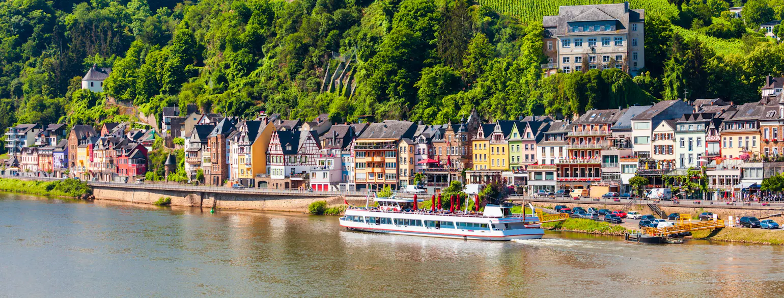 Colorful riverside European houses below a green hillside, tour boat cruising on the river