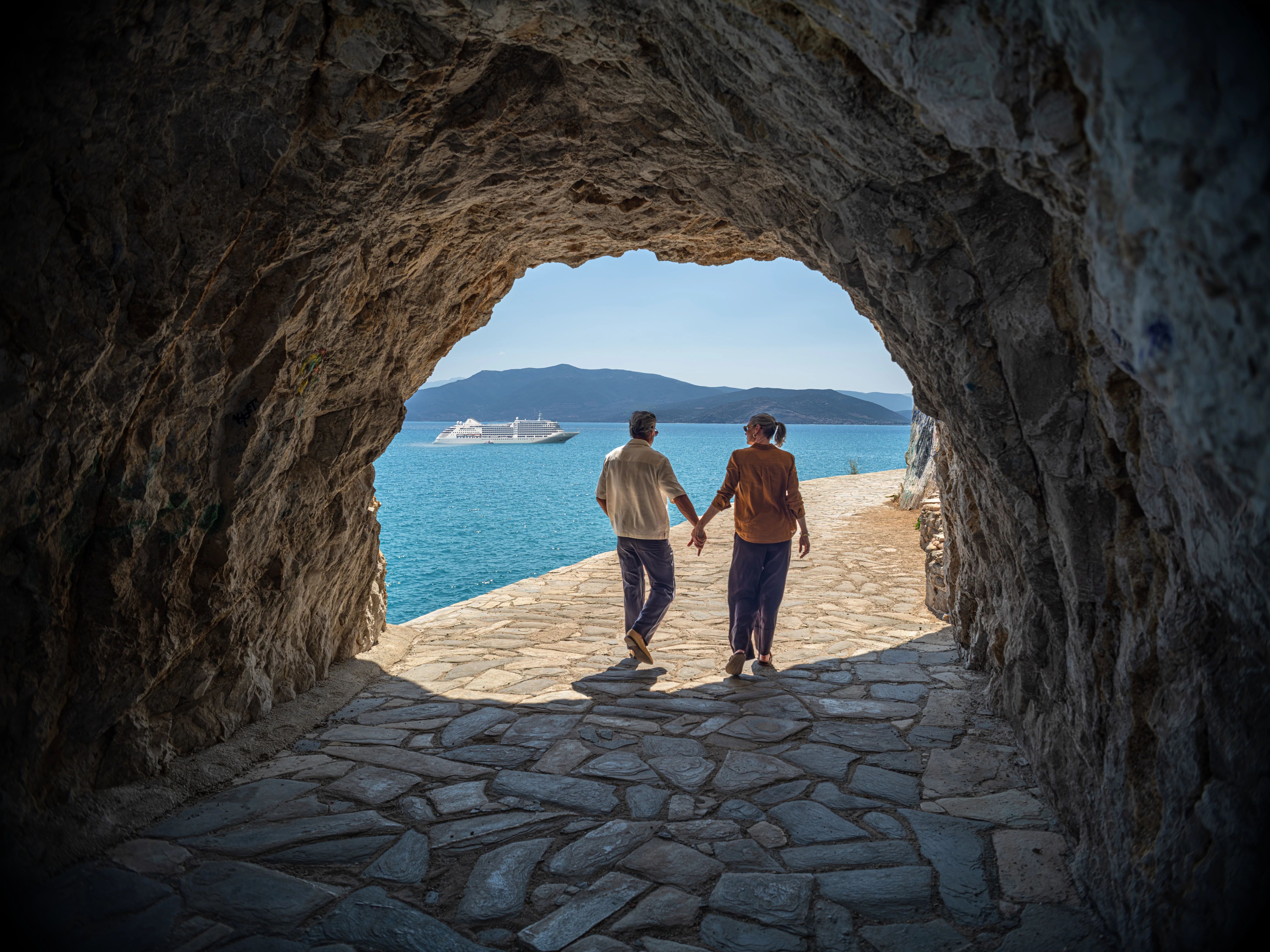 Couple holding hands walking from cave onto seaside path with cruise ship and mountains