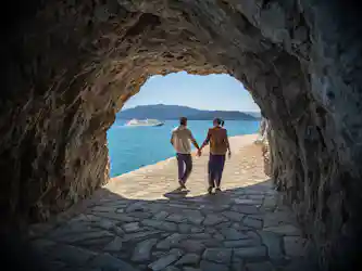 Couple holding hands walking from cave onto seaside path with cruise ship and mountains