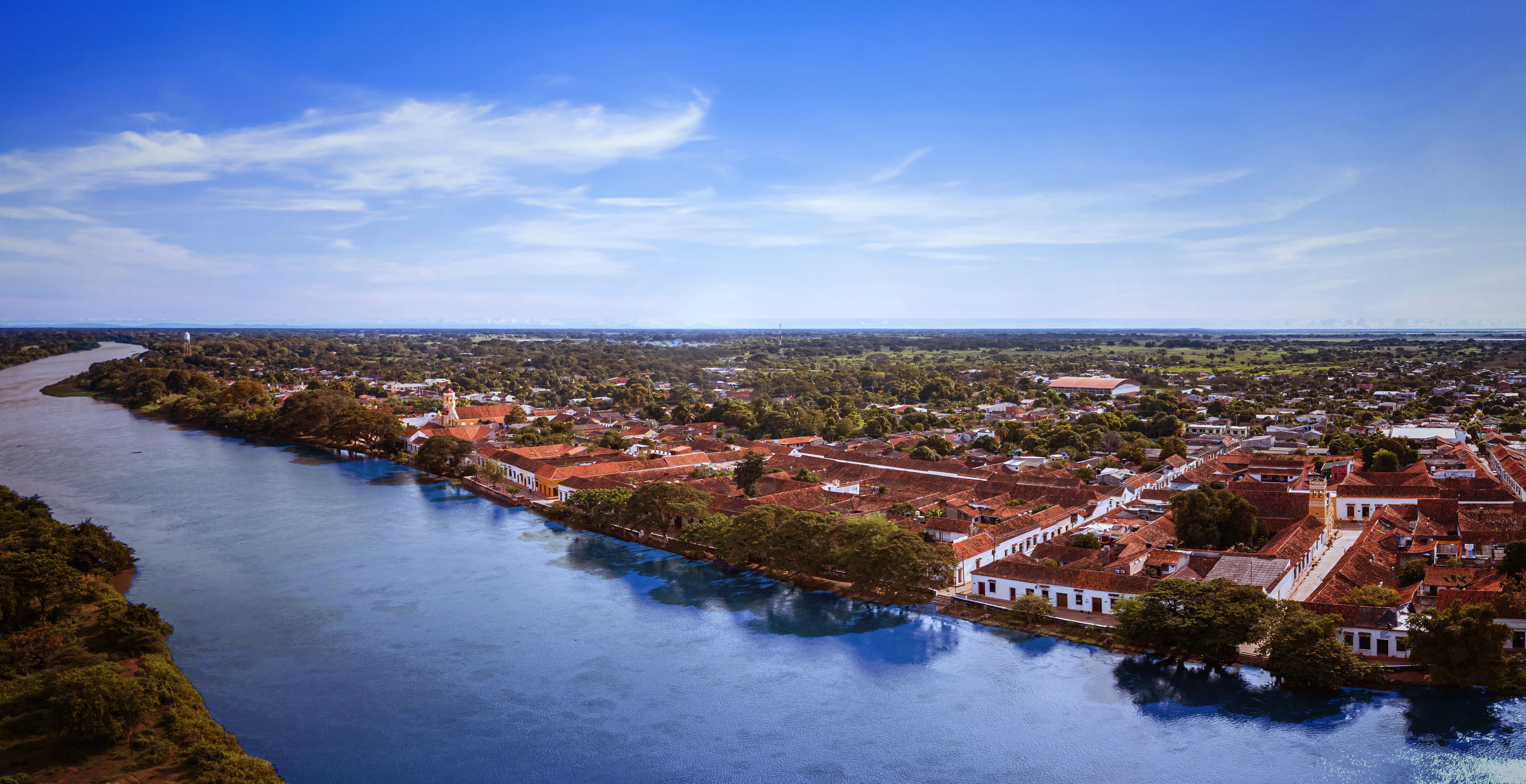Aerial panorama of colonial riverside town with red-tiled roofs, riverside trees, and church tower