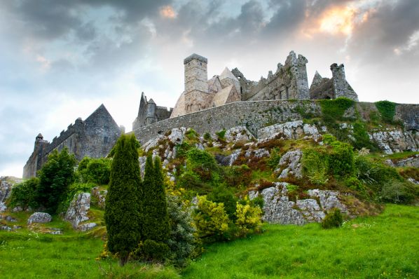 Medieval stone castle ruins atop a rocky green hill with trees beneath a dramatic cloudy sky