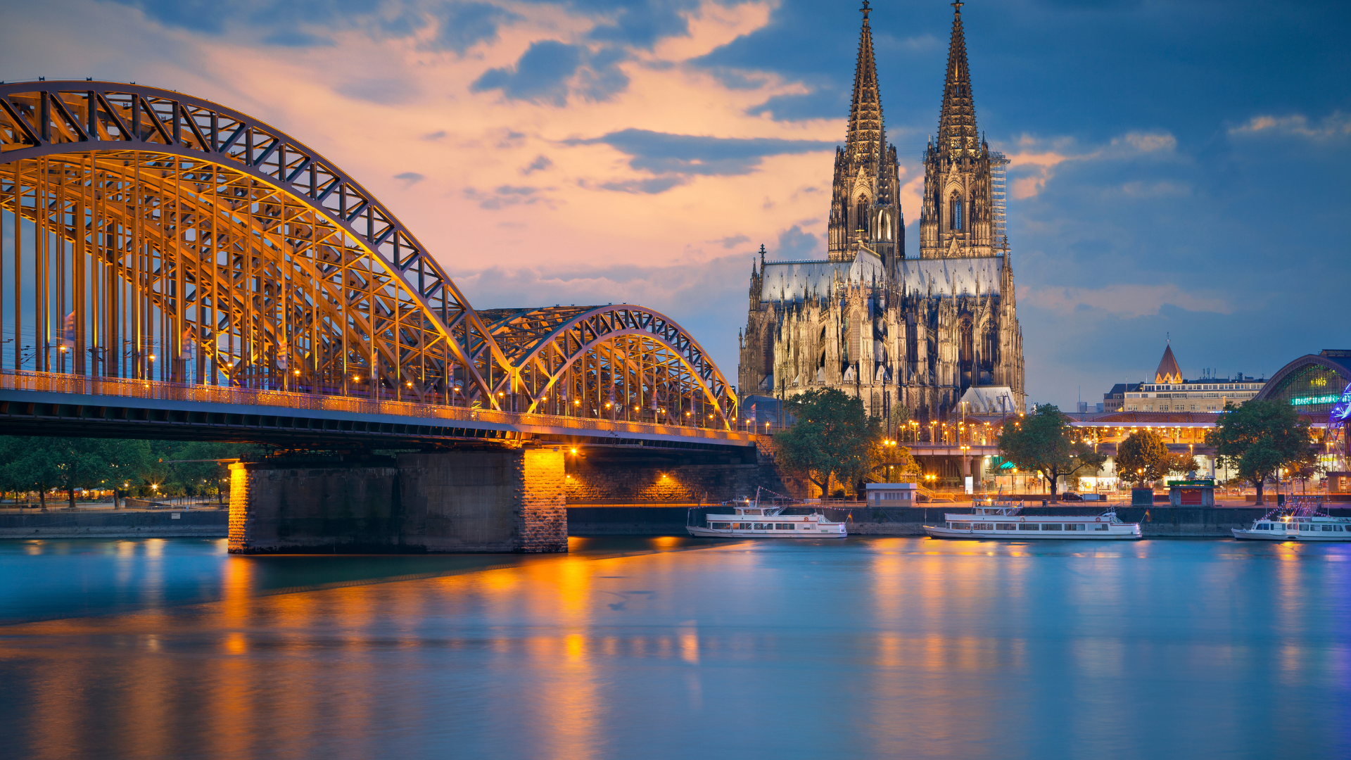 Hohenzollern Bridge arches and illuminated Cologne Cathedral reflected on the Rhine, boats at dusk