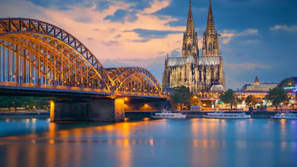 Hohenzollern Bridge arches and illuminated Cologne Cathedral reflected on the Rhine, boats at dusk