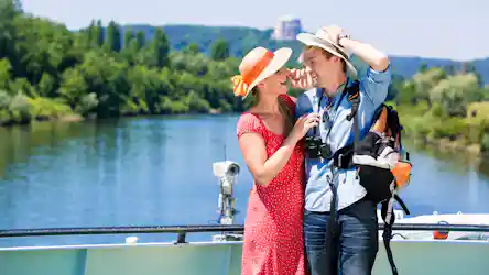 Couple in sun hats laughing on a boat deck by a river; woman in red dress, man with backpack