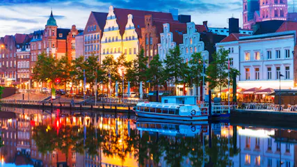 Evening canal with moored tour boat, illuminated historic gabled buildings, trees and waterside cafes reflected