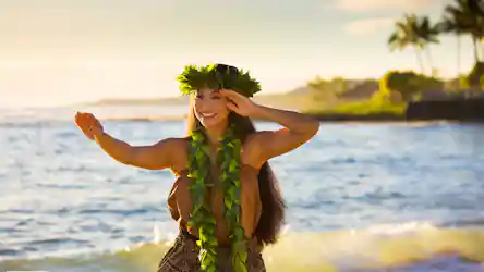 Smiling hula dancer in leaf lei and crown performing on a sunlit beach with ocean waves
