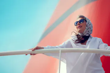 Woman in patterned headscarf and sunglasses leaning on boat railing, gazing outward