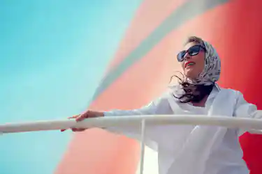 Woman in patterned headscarf and sunglasses leaning on boat railing, gazing outward
