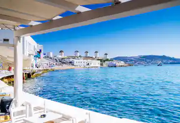 Café terrace overlooking turquoise Aegean, white Cycladic houses and Mykonos windmills