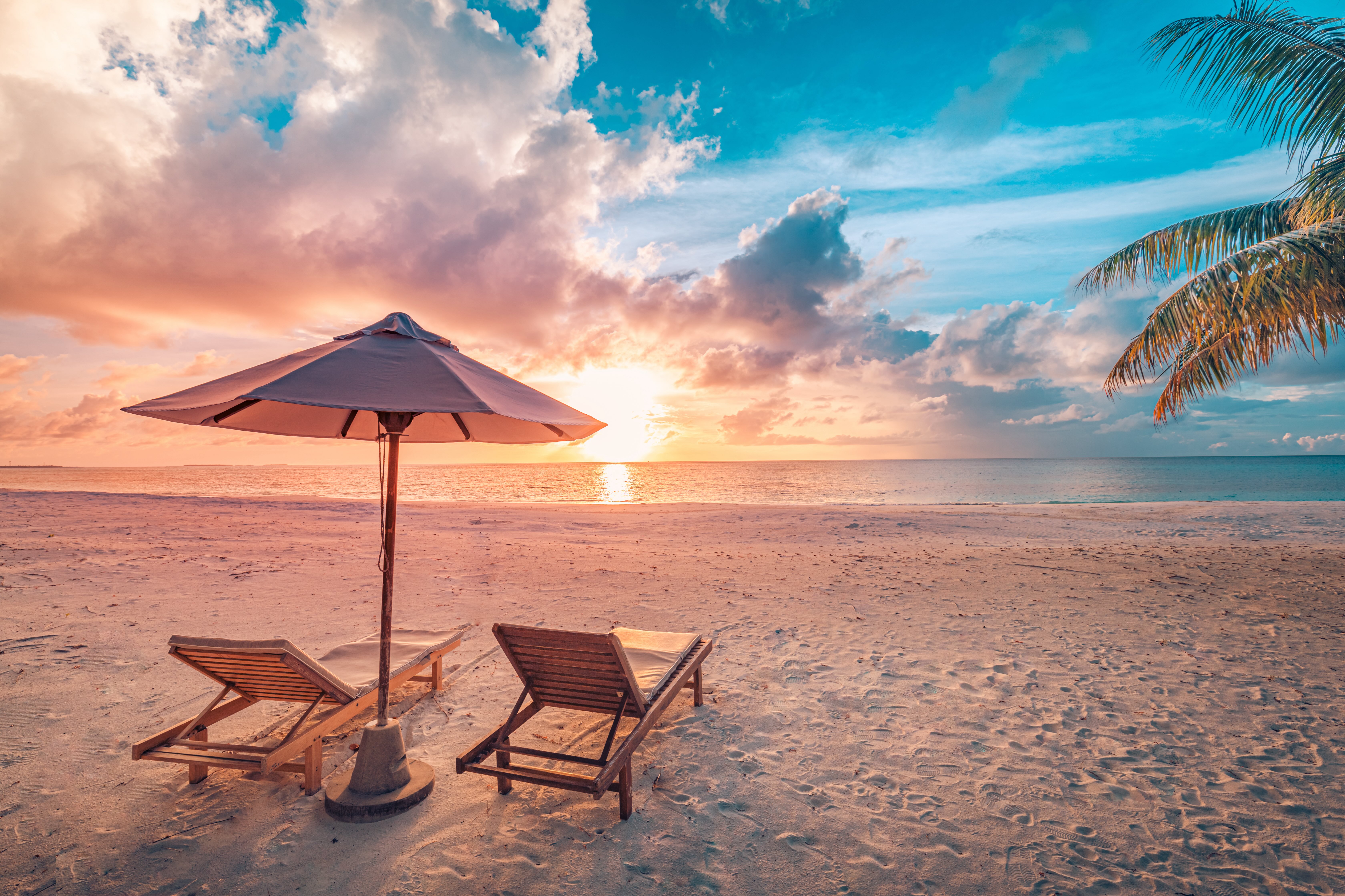 Sunset beach with two wooden lounge chairs under umbrella, palm fronds, sun reflecting on ocean.