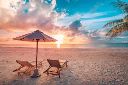 Sunset beach with two wooden lounge chairs under umbrella, palm fronds, sun reflecting on ocean.