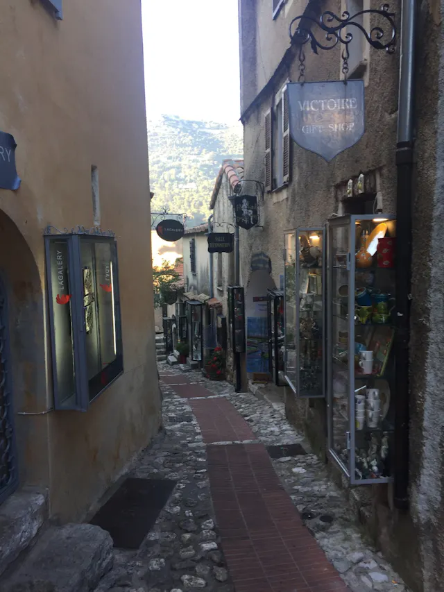 Narrow cobblestone alley with gift shop displays, hanging signs and distant hills.