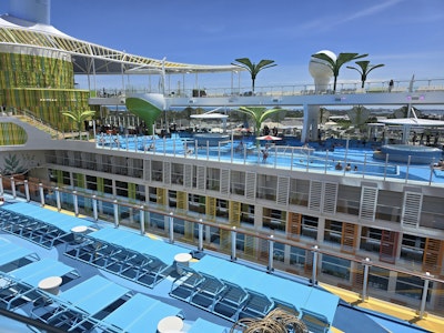 Multi-level cruise ship pool deck with turquoise loungers, pools, green palm-like canopies