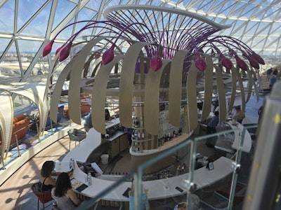 Curved white bar under a glass dome with wooden ribs and magenta hanging lights on a cruise ship