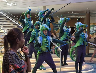 Six performers in blue-green sea creature costumes perform with staffs on cruise ship lobby stairs