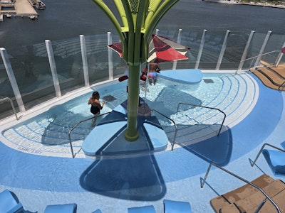 Shallow ship deck splash pool with green canopy, red umbrella, wading swimmers, glass railing