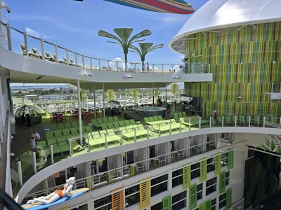 Multi-deck cruise ship sun deck with green lounge chairs, palm-shaped shades, colorful slatted wall.