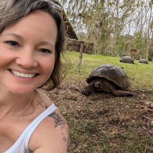 Smiling selfie showing shoulder tattoo beside a giant tortoise on grass, two more behind