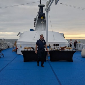 Crew member standing between buffet tables on blue cruise ship deck at sunset