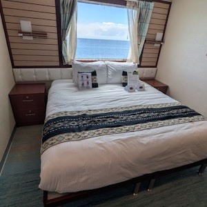 Bed in cruise cabin with white linens, patterned runner, pillows, nightstands, ocean visible