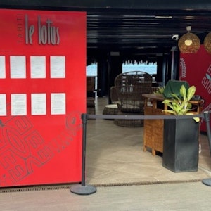 Restaurant entrance with red 'le lotus' menu board, roped host desk, wicker seating, lagoon view