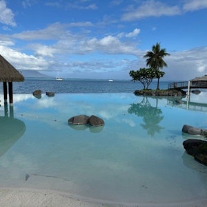 Infinity pool at a tropical resort blending into the ocean, sandy shore, palm tree, rocks, sailboat.
