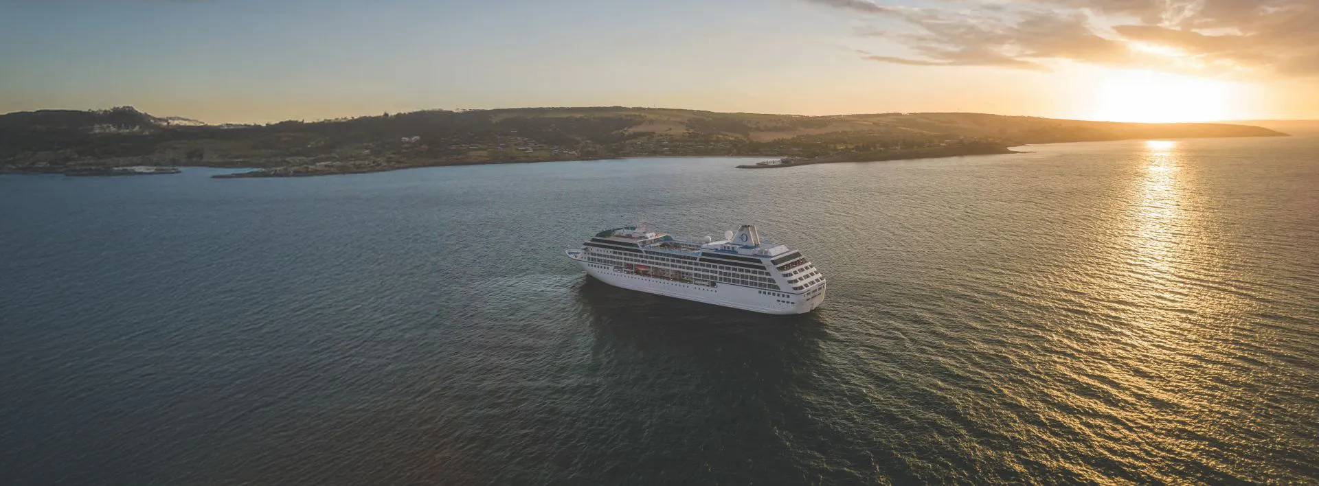 White cruise ship off a hilly coastline at sunset with golden reflections on the sea