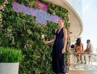 Woman ringing brass bell by a leafy green wall with neon Sanctuary Club sign