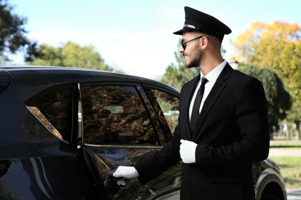 Chauffeur in black suit, white gloves, cap and sunglasses opening a black SUV door by park trees