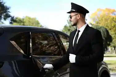 Chauffeur in black suit, white gloves, cap and sunglasses opening a black SUV door by park trees
