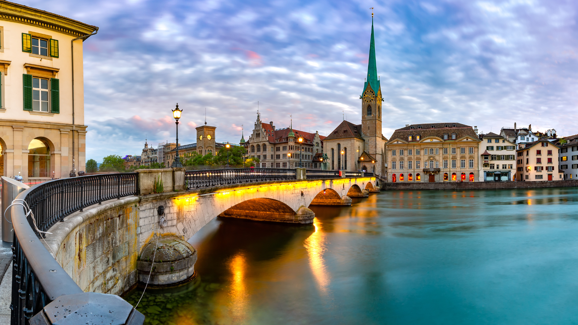 Arched stone bridge lit at dusk over river, Gothic church spire and historic buildings