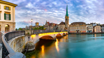 Arched stone bridge lit at dusk over river, Gothic church spire and historic buildings