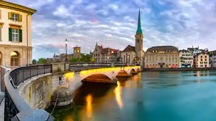 Arched stone bridge lit at dusk over river, Gothic church spire and historic buildings