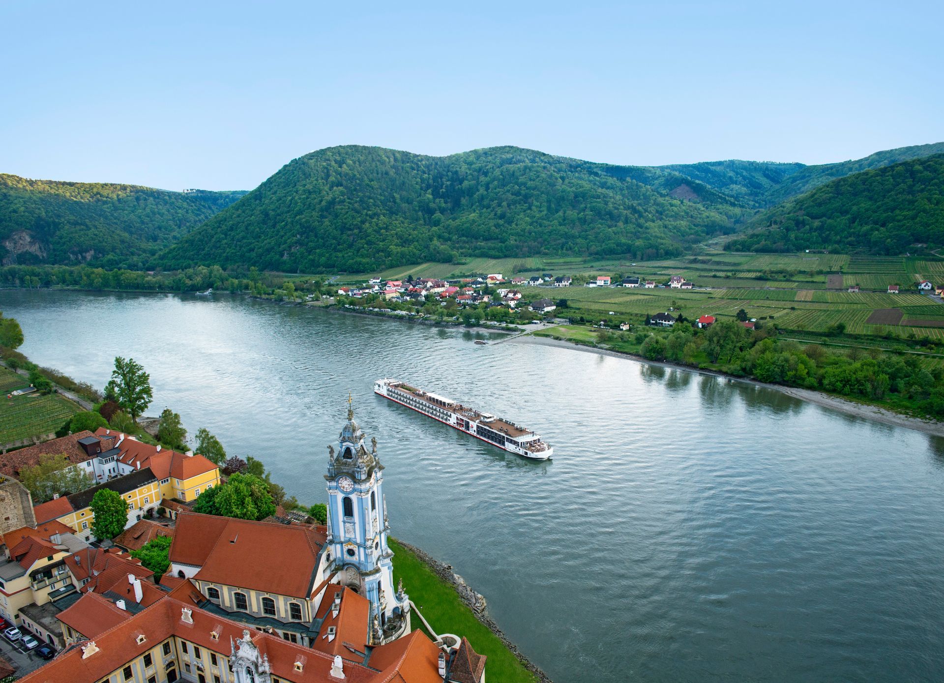 Cruise ship on river past baroque church tower and red-roof village with vineyard-covered hills