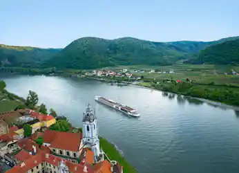 Cruise ship on river past baroque church tower and red-roof village with vineyard-covered hills