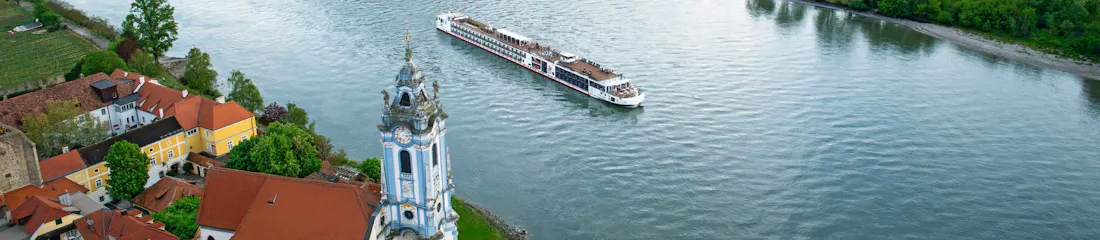 Cruise ship on river past baroque church tower and red-roof village with vineyard-covered hills