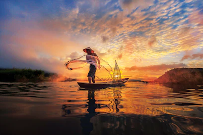 Fisherman in conical hat casting a circular net from a boat at sunset, water reflection